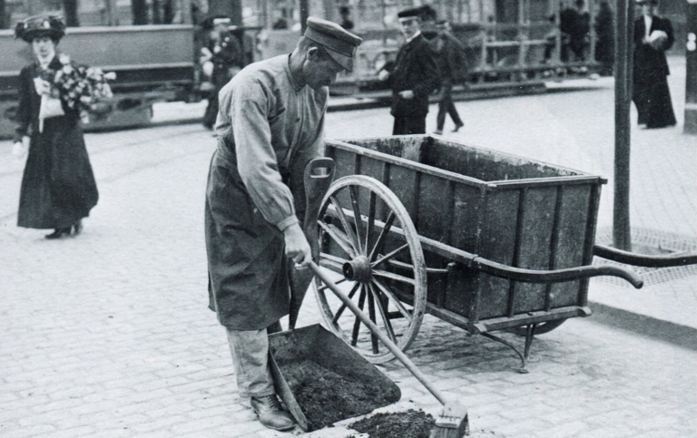svartvitt foto av man i gammaldags kläder som sopar upp avfall på gatan.