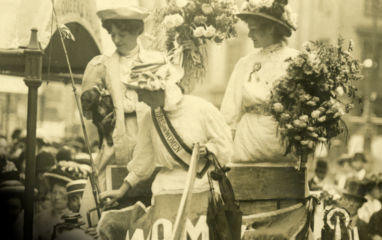Svartvit bild med tre kvinnor i hatt, den ena med ett band över axels med texten "votes for women"
